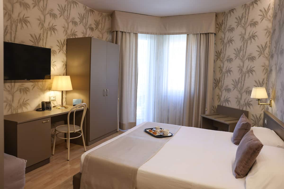 Une chambre d’hôtel avec un lit, un bureau, une chaise, une télévision et une fenêtre avec des rideaux.