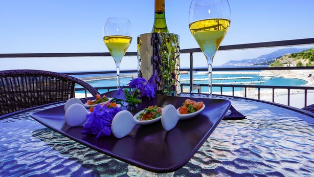 Een tafel met een fles wijn, twee wijnglazen en een bord met eten en bloemen op een balkon met uitzicht op een strand.