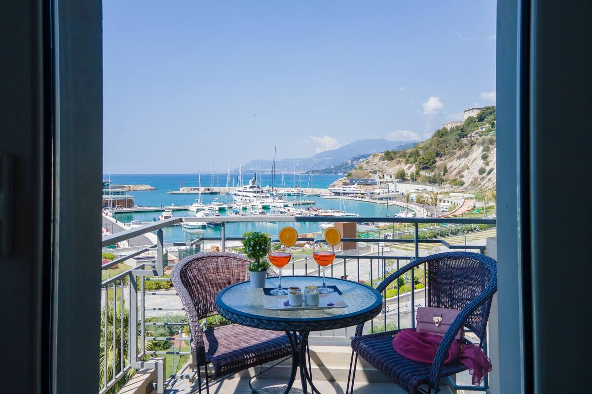 Un balcon agrémenté de fauteuils et d’une table en osier, offrant une vue sur une marina avec des bateaux et un paysage vallonné.