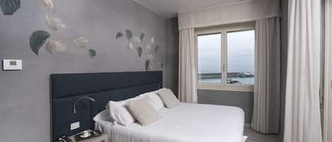 A modern hotel room with a large bed, a bedside table with a lamp, and a view of the sea through a window.