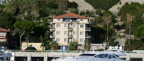 A marina with a white yacht docked in the foreground. In the background, there is a multi-story building with balconies and a hillside with vegetation.