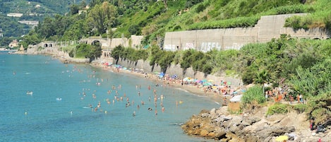 A coastal beach with clear blue water, rocky shoreline, and a stone wall with arches.