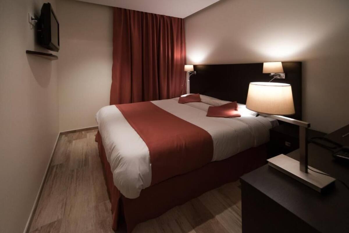 Habitación de hotel con una cama grande, dos lámparas de noche, un televisor de pantalla plana montado en la pared y suelo de madera.