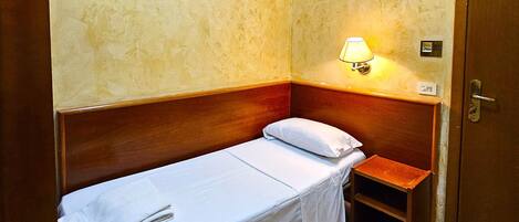 A small, single-bed room with a wooden headboard, a bedside table, a wall-mounted air conditioner, and a door.