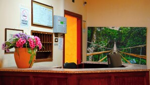 A reception desk with a flower vase, a framed notice, a digital display, and a poster of a rope bridge in a forest. - Ferrari (Rome)