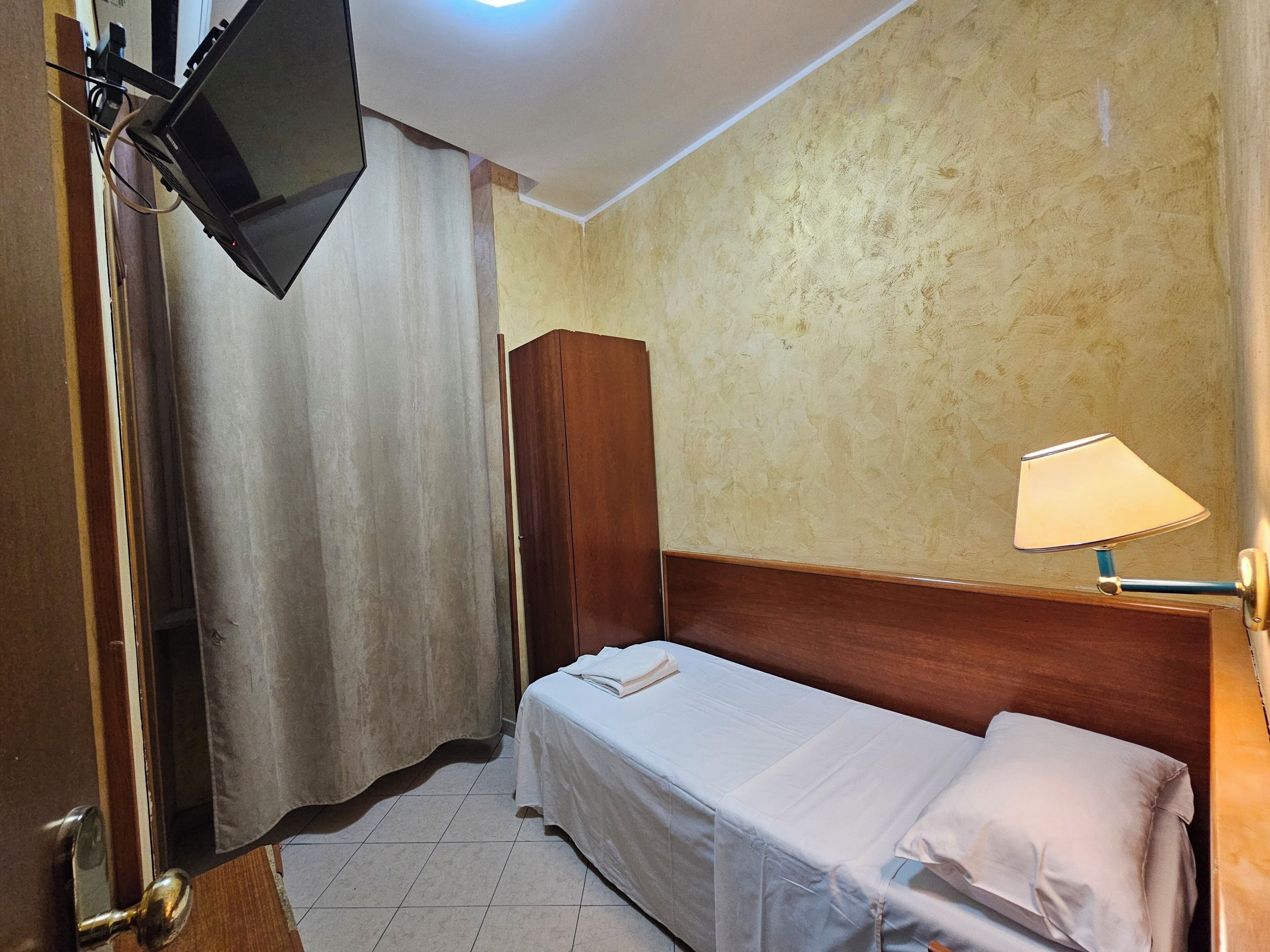 Habitación de hotel con cama, armario de madera, televisor montado en la pared y lámpara en una mesita de noche.