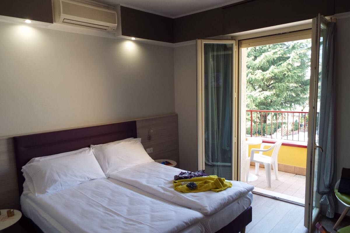 Une chambre d’hôtel avec un lit, une table de chevet, une fenêtre donnant sur un espace verdoyant et un balcon avec une chaise.
