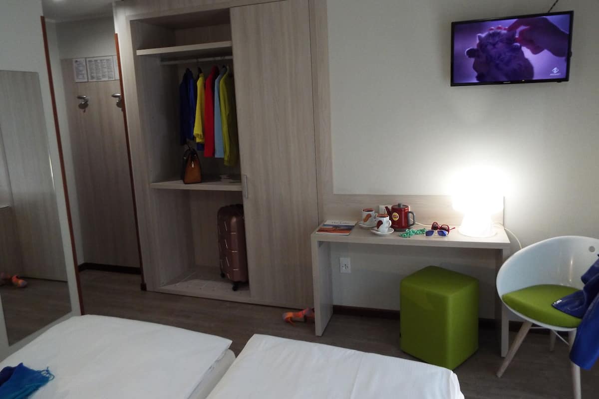 Une chambre d’hôtel avec un lit, un bureau doté d’une lampe, un pouf vert, une chaise, une armoire et un téléviseur fixé au mur.