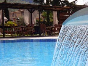 A pool area with a water feature, lounge chairs, and a covered seating area. - Imperial (Garda)