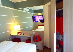 A hotel room with a bed, a desk with a lamp, a television on the wall, a red handbag, and a closet with clothes and shoes. - Imperial (Garda)