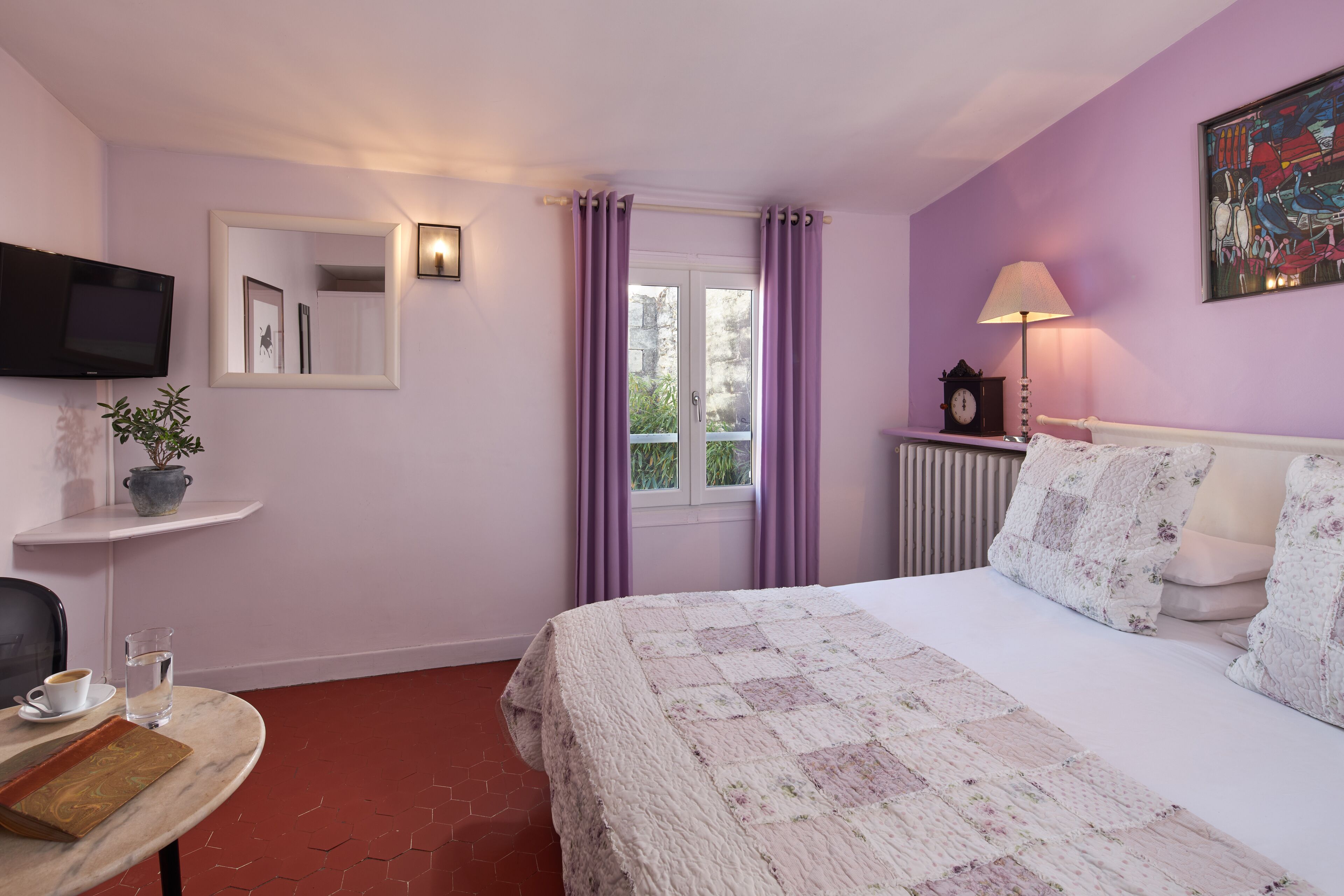 A bedroom with a bed, a TV, a window with curtains, a bedside table with a lamp, and a wall-mounted painting.