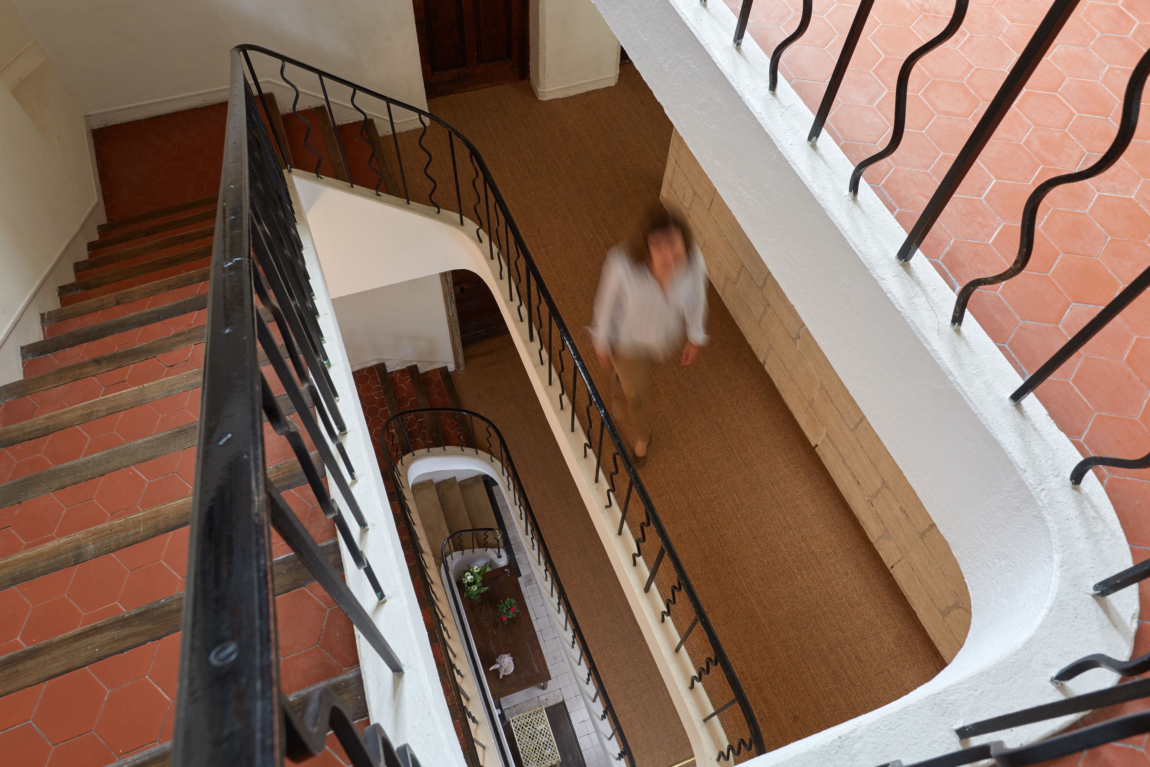 Un intérieur à plusieurs niveaux avec un escalier, un coin repas comprenant une table et des chaises, et un mur blanc orné d’un motif hexagonal.