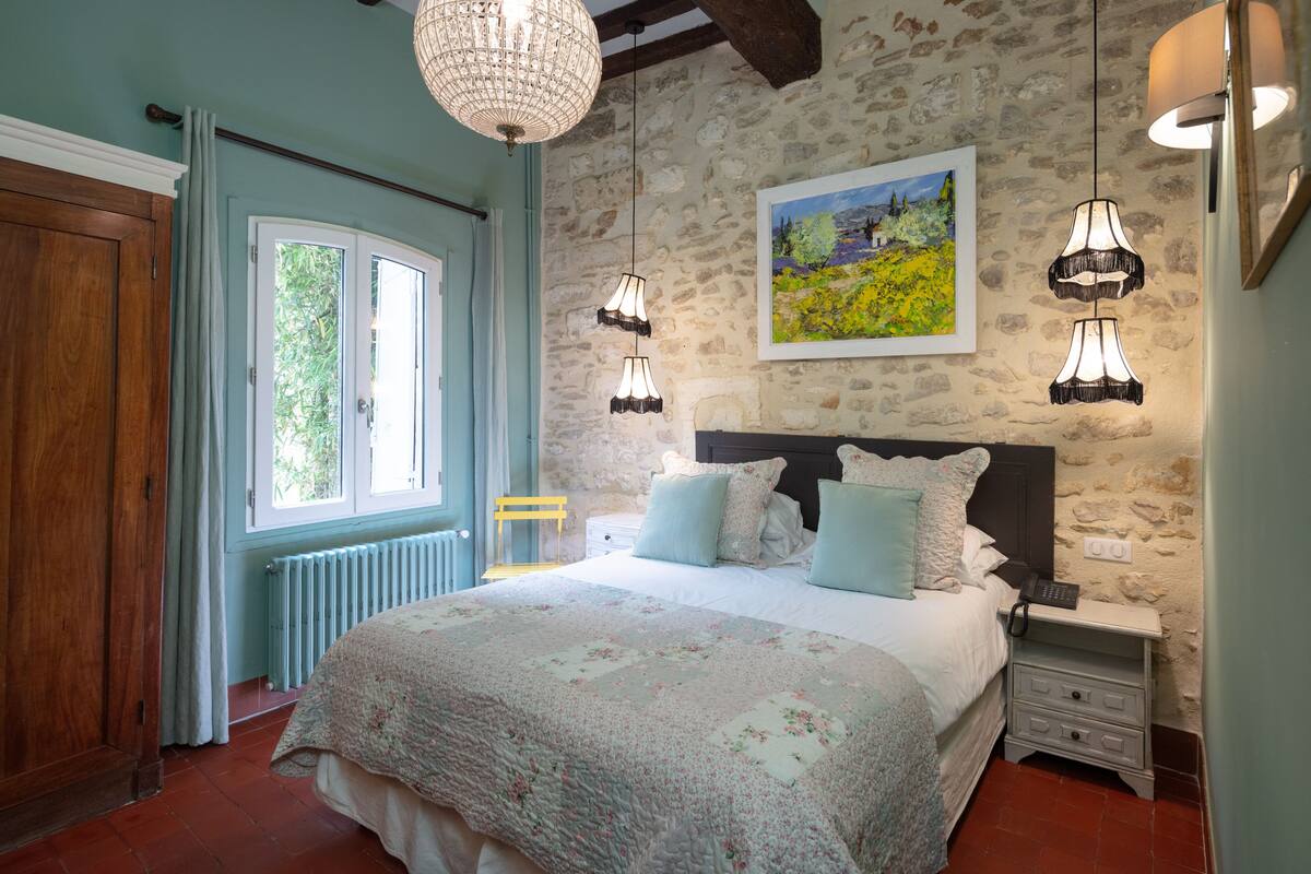 Une chambre avec un mur en pierre, un lit recouvert d’une courtepointe à motifs floraux et deux lampes suspendues.