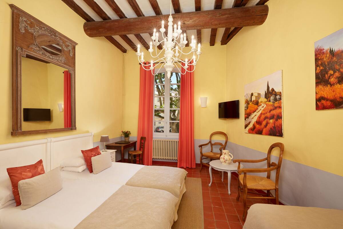 Une chambre d’hôtel avec un grand lit, deux fauteuils, une petite table, un lustre, un miroir et un tableau au mur.