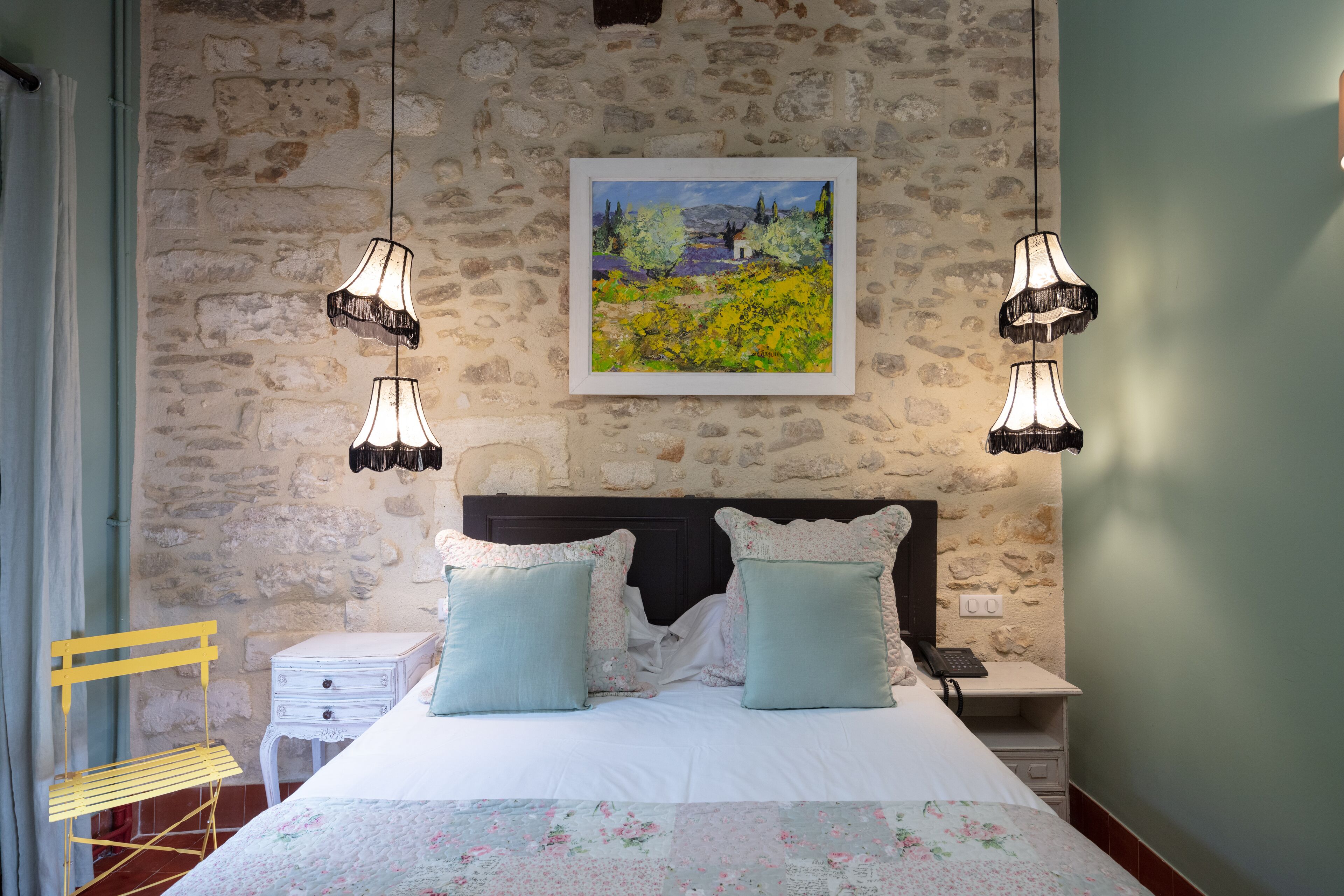 Un dormitorio con pared de piedra, cama con ropa de cama floral, dos mesitas de noche y lámparas colgantes.