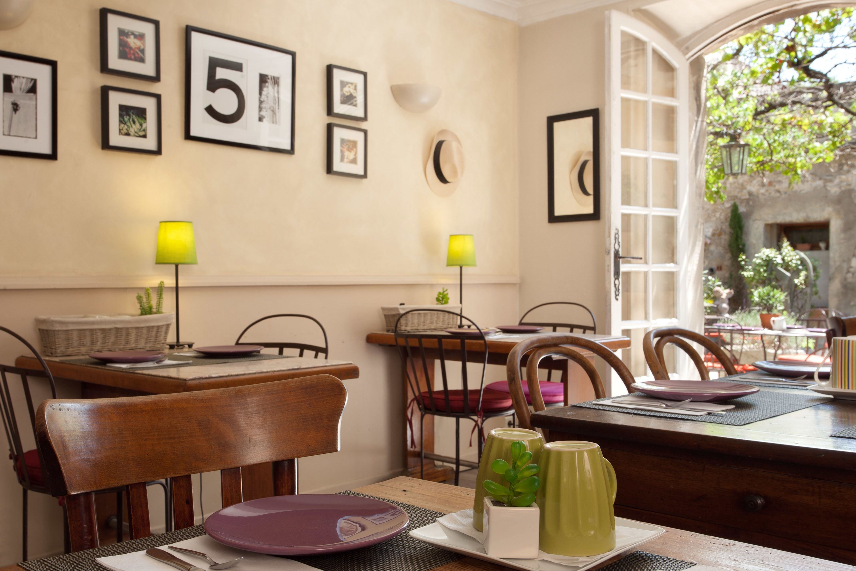 A dining area with wooden tables and chairs, framed pictures on the wall, and a view of the outdoor garden.