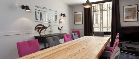 A modern dining area with a long wooden table, pink and gray upholstered chairs, black pendant lights, and a decorative metal sculpture.