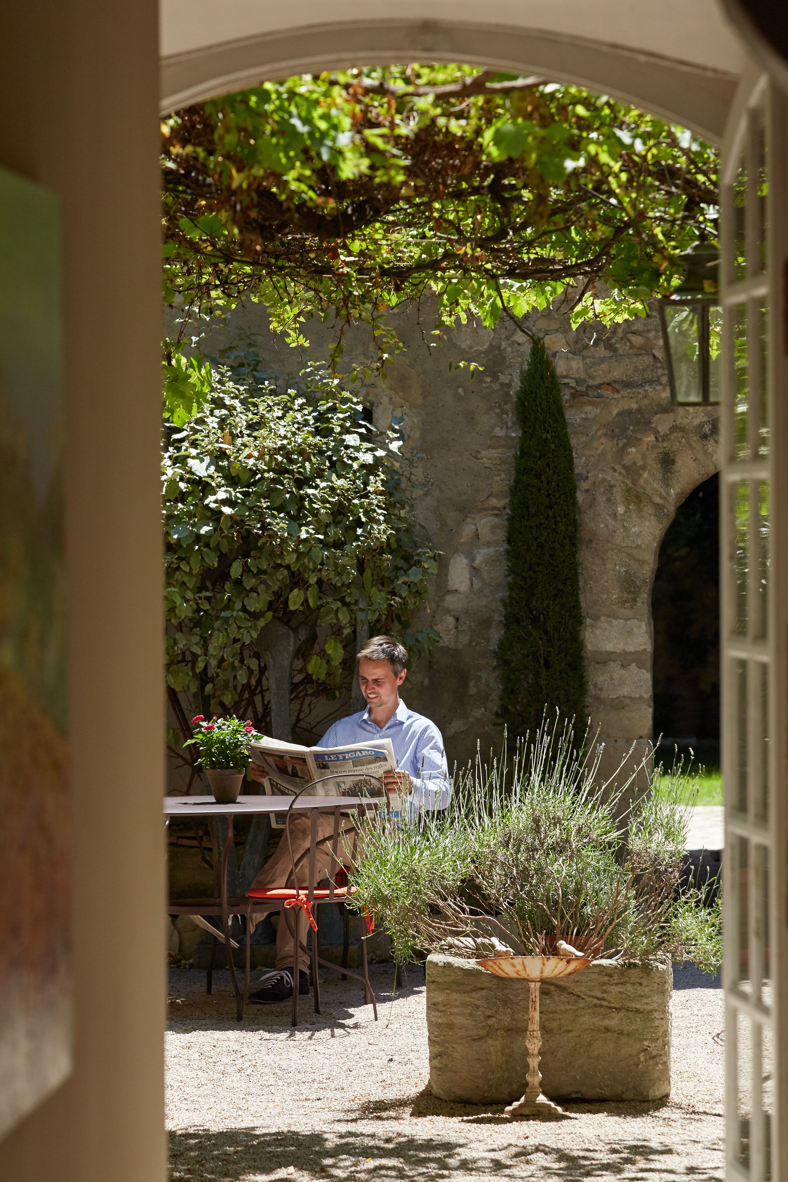 A man reading a newspaper at a table in a garden with stone walls and potted plants.
