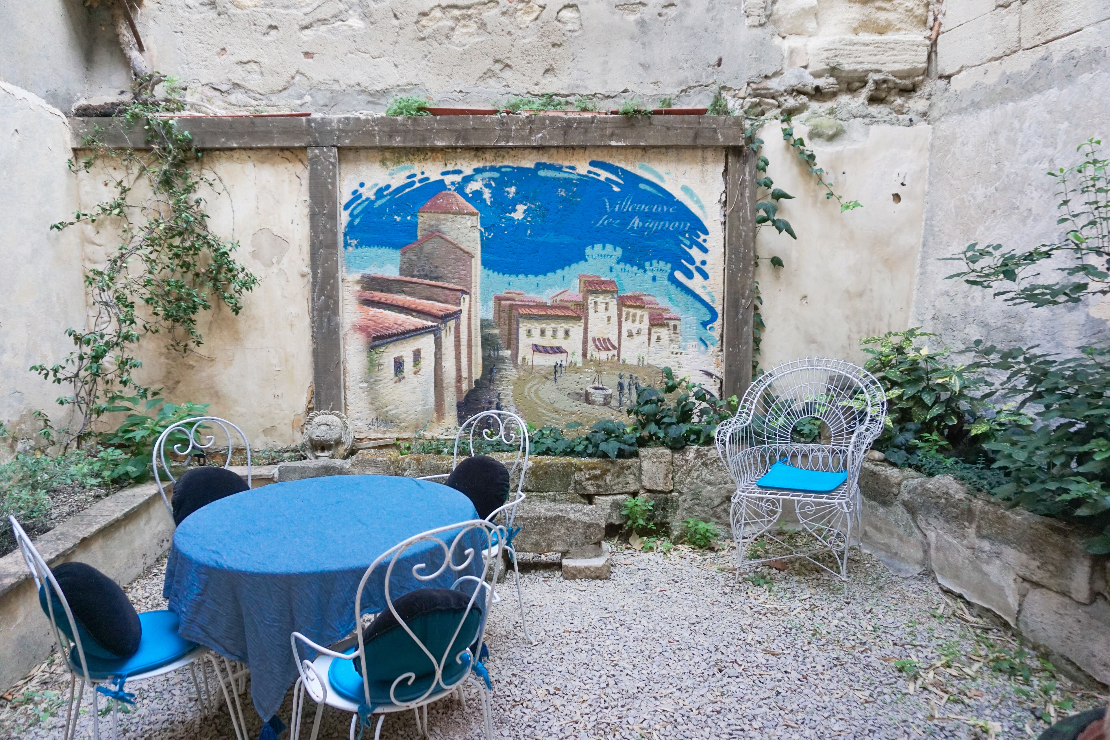A courtyard with a round table and chairs, a mural on the wall, and a stone wall.