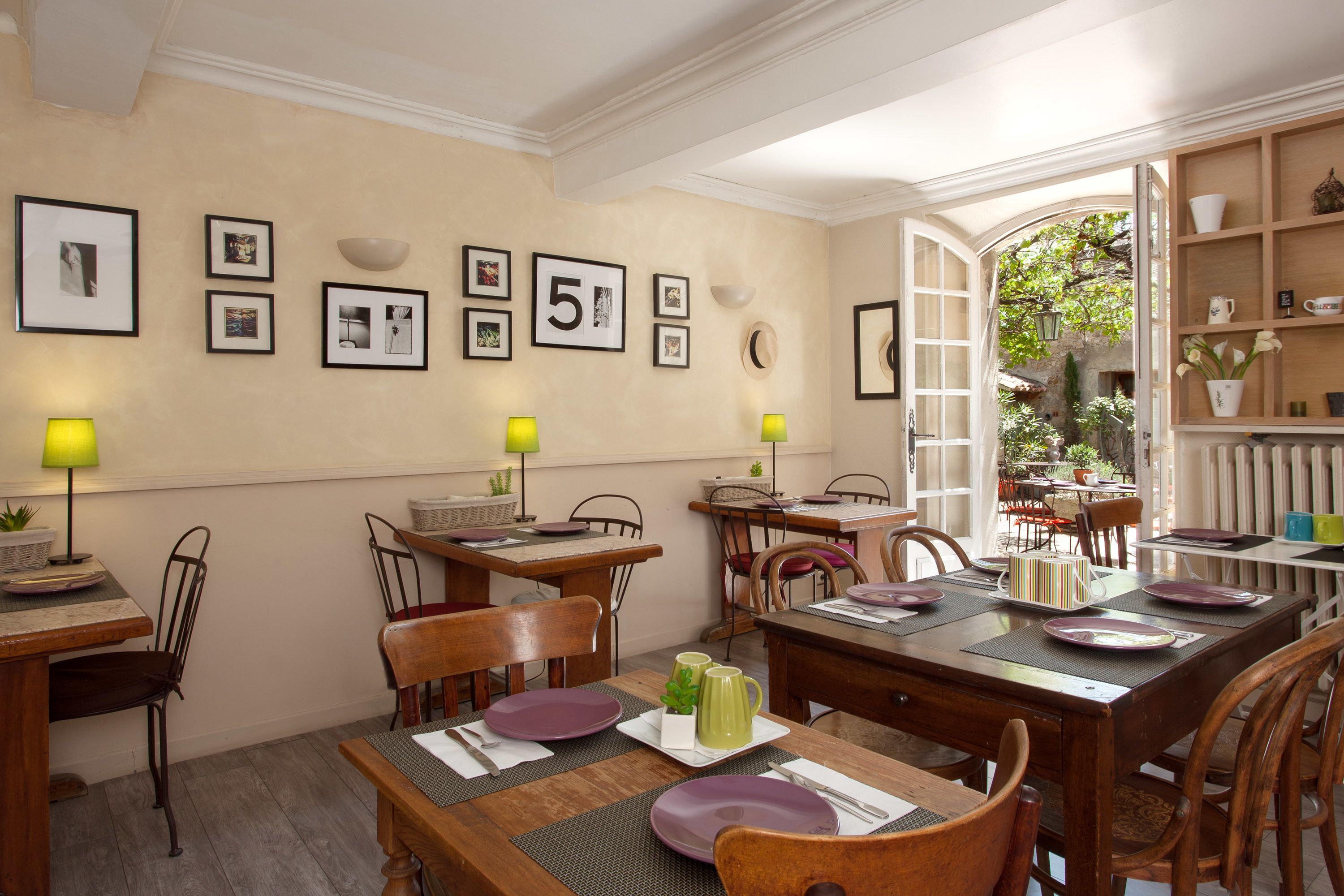 A dining area with wooden tables and chairs, framed pictures on the wall, and a view of the garden through an open door.
