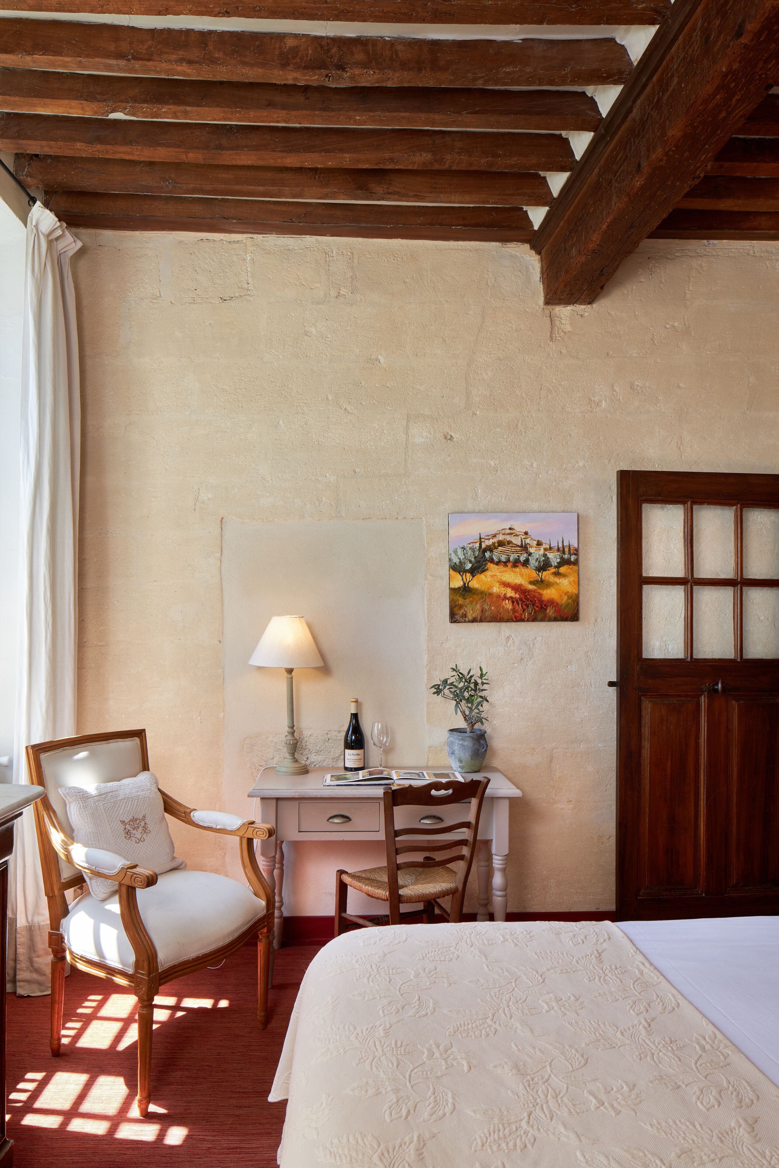 A bedroom with a bed, a desk with a lamp, a chair, a wooden door, and a painting on the wall.
