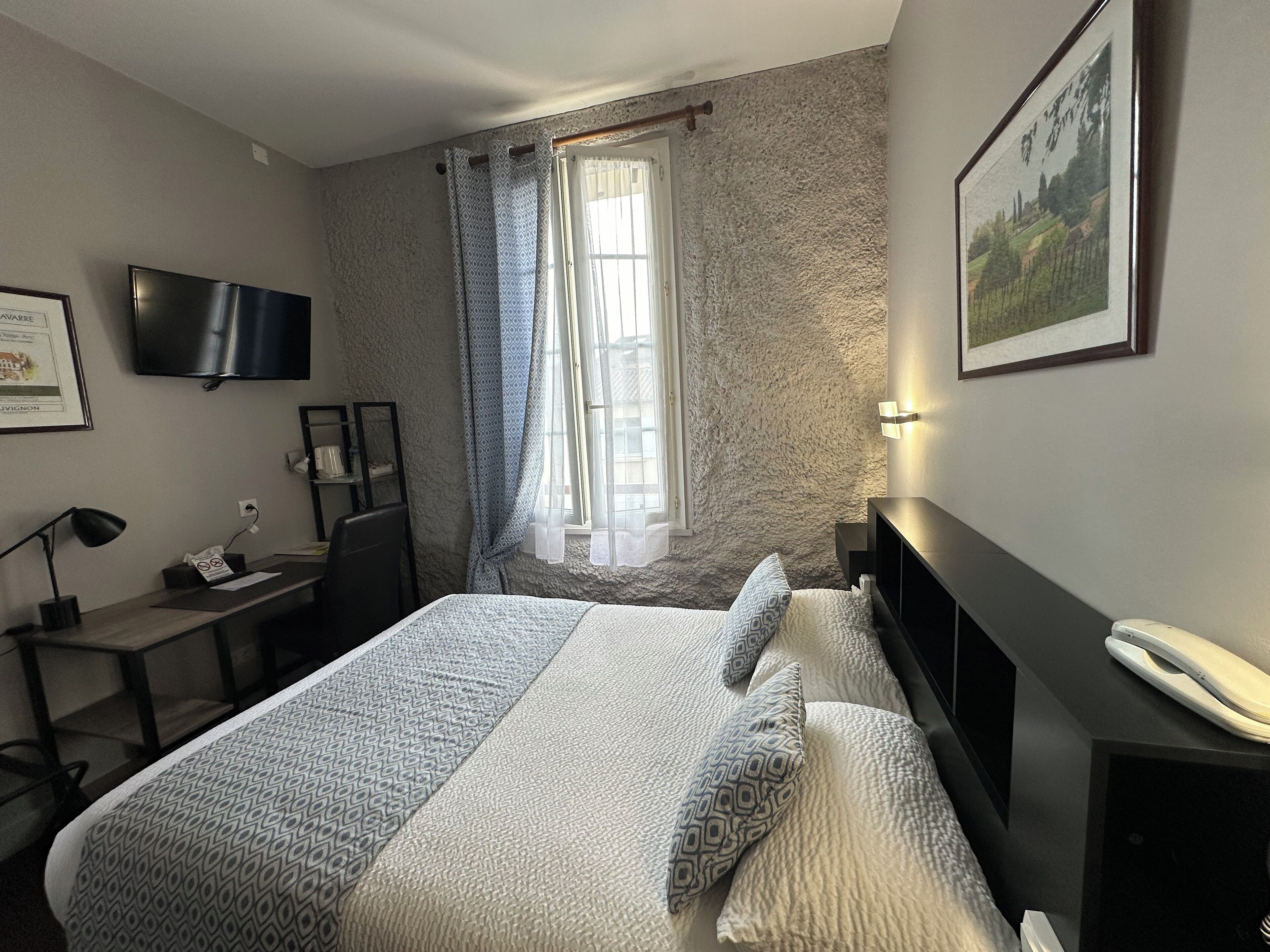 Une chambre d’hôtel comprenant un lit, un bureau, une chaise, une télévision, une fenêtre avec des rideaux et un tableau encadré au mur.