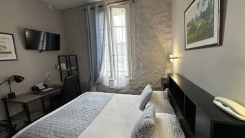 Habitación de hotel con cama, escritorio, silla, televisor, ventana con cortinas e imagen enmarcadas en la pared.