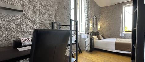 A room with a wooden floor, a desk with a chair, a bed with pillows, and a stone wall.