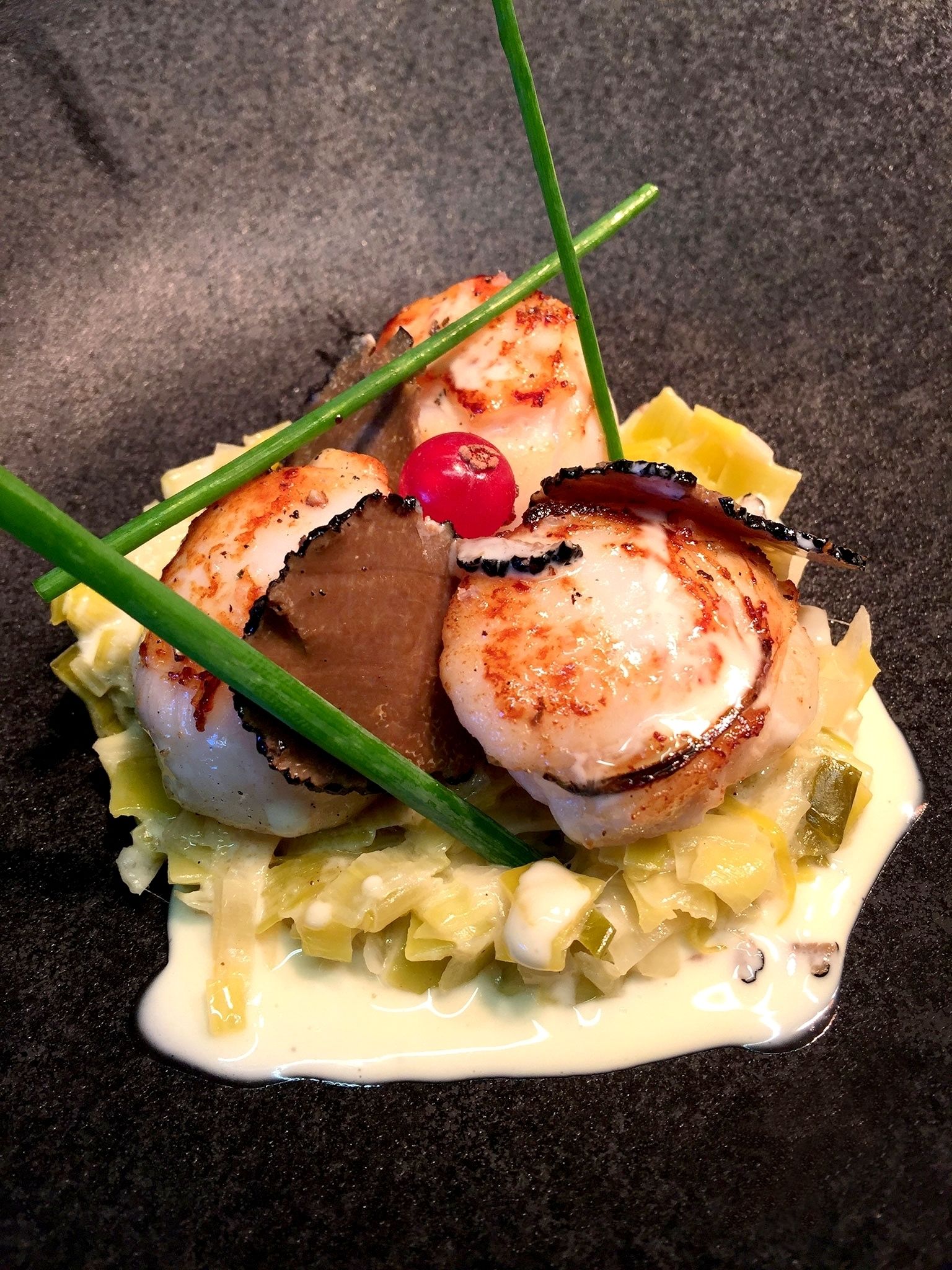 A dish with seared scallops, mushrooms, and a cherry on a bed of greens and vegetables.