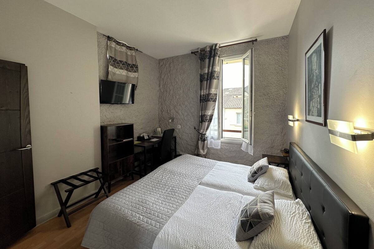 Quarto com cama, escrivaninha, cadeira, televisão e janela com cortinas.