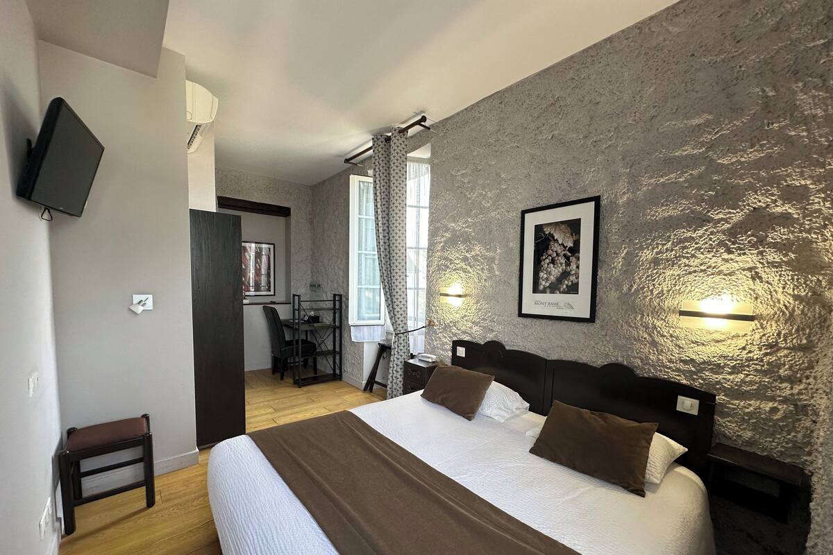 Quarto com uma cama grande, uma escrivaninha e uma cadeira. Há uma televisão na parede e um quadro emoldurado.