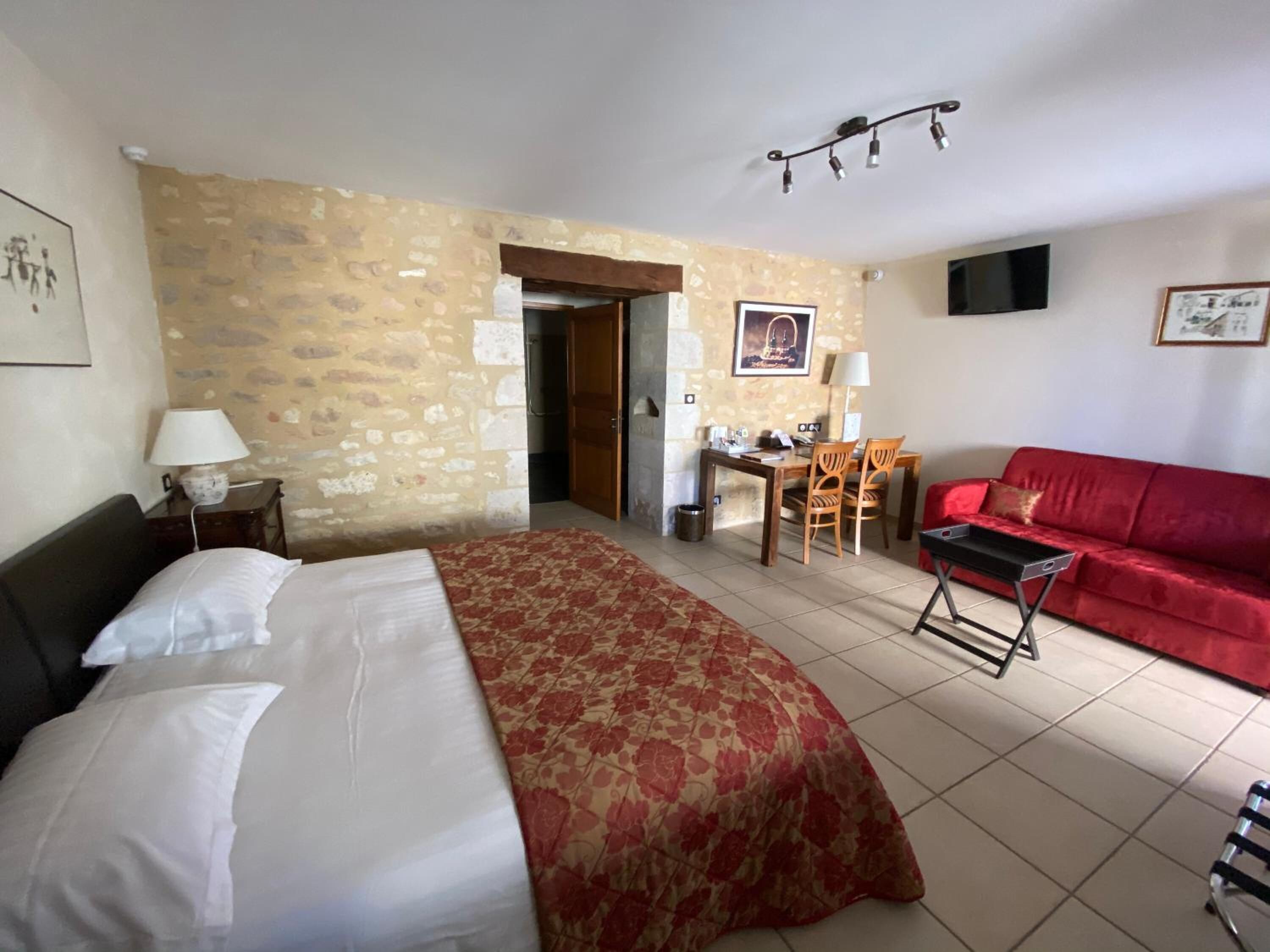 A hotel room with a bed, a red sofa, a desk with chairs, and a TV on the wall.