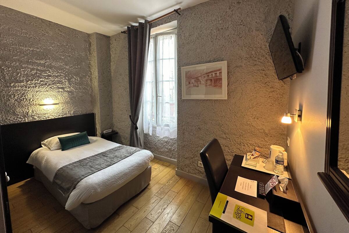 Quarto de hotel com cama, escrivaninha com cadeira, janela com cortinas, televisão fixada na parede e luminária.