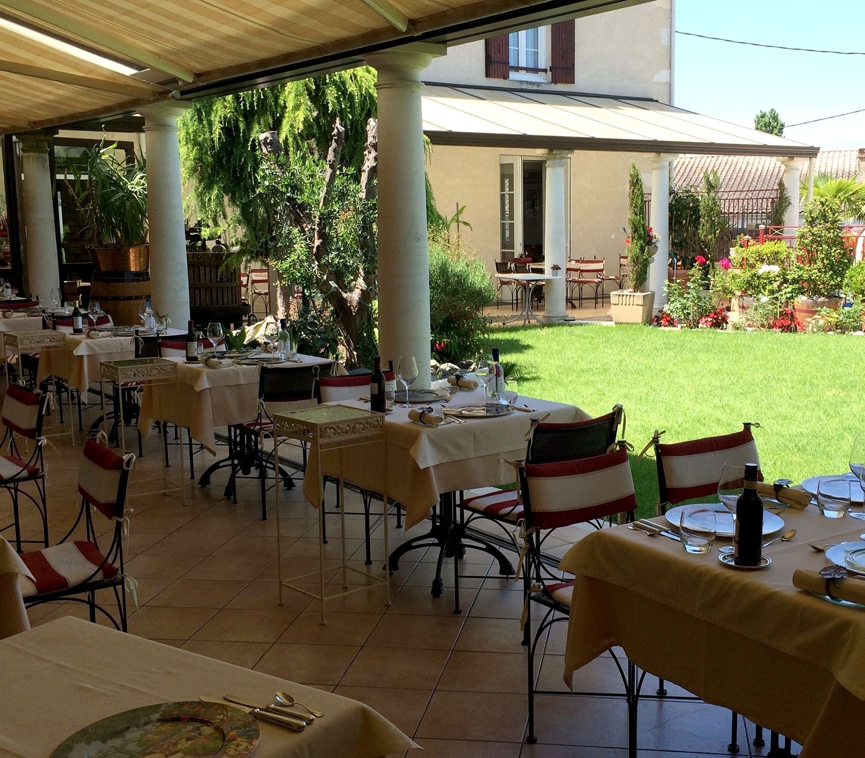 Un área de comedor al aire libre con mesas y sillas, rodeada de vegetación y un edificio al fondo.