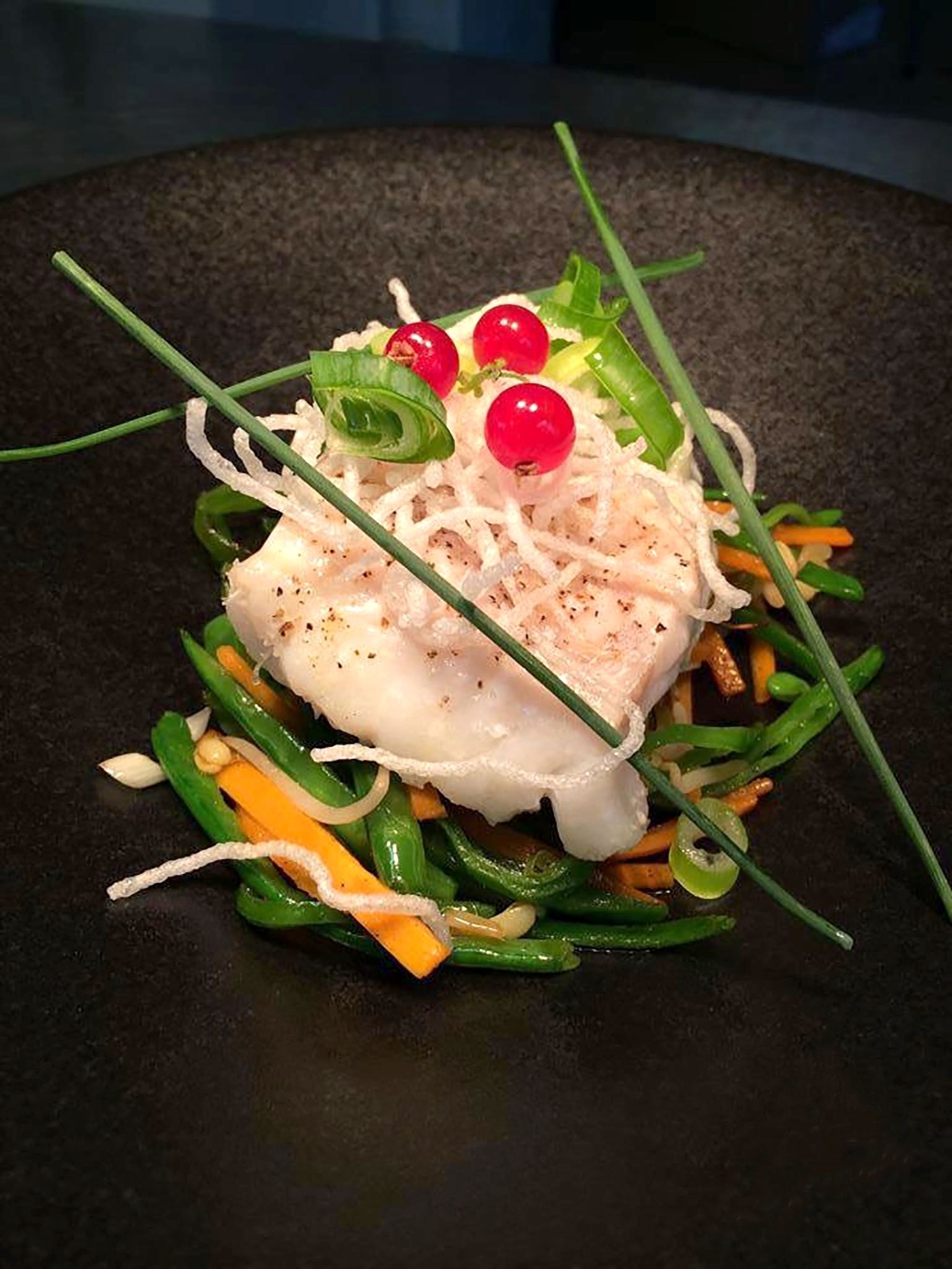 A dish with white fish, green vegetables, and red berries on a dark plate.