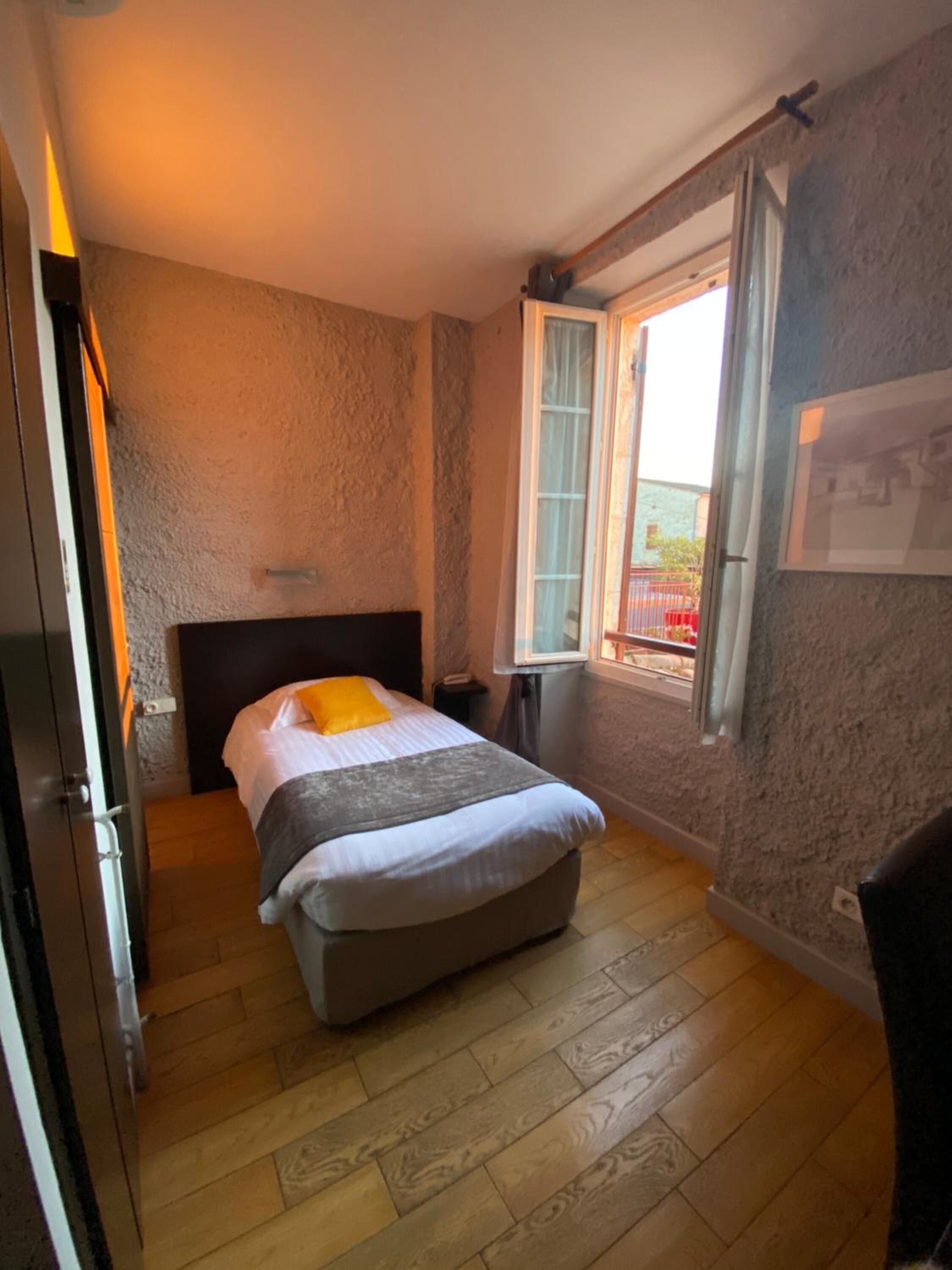 A small, cozy bedroom with a single bed, a wooden floor, a window with curtains, and a door.