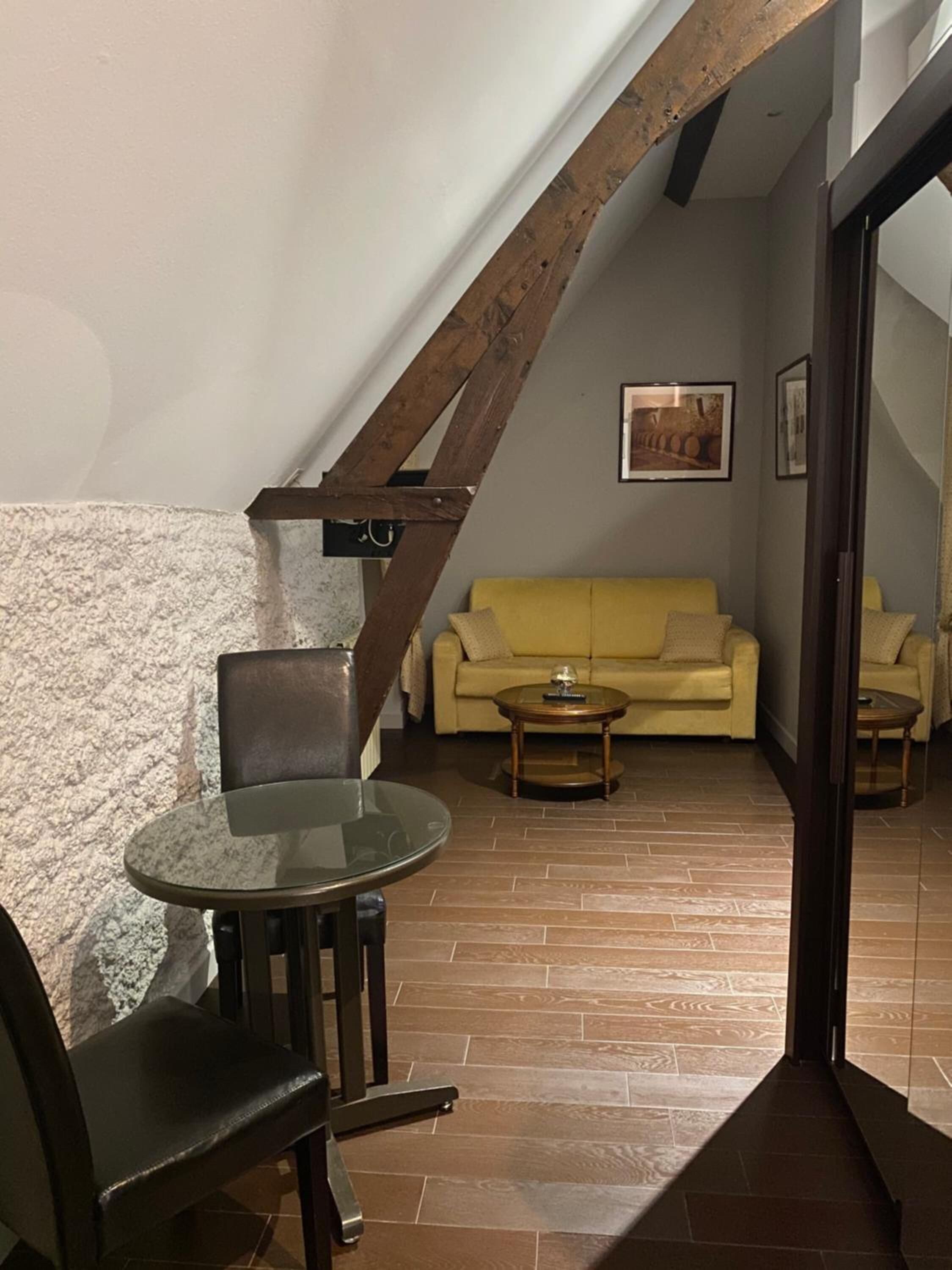 Un espacio de sala acogedor con un sofá amarillo, una mesa de vidrio y una silla de madera. Una escalera con vigas a la vista conduce a otra habitación.