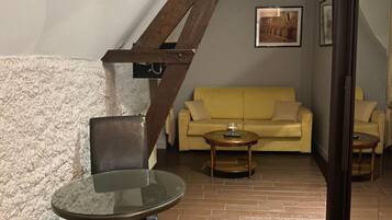Un espacio de sala acogedor con un sofá amarillo, una mesa de vidrio y una silla de madera. Una escalera con vigas a la vista conduce a otra habitación.