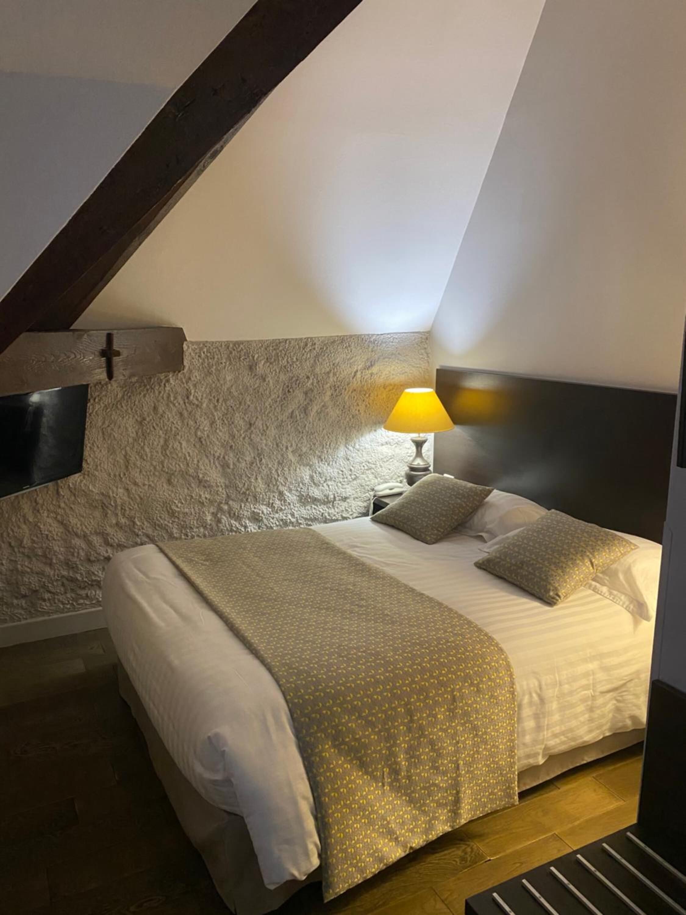 Ein Schlafzimmer mit schrägen Wänden, einem Bett mit gemustertem Überwurf, Kissen, einer Nachttischlampe und Holzboden.