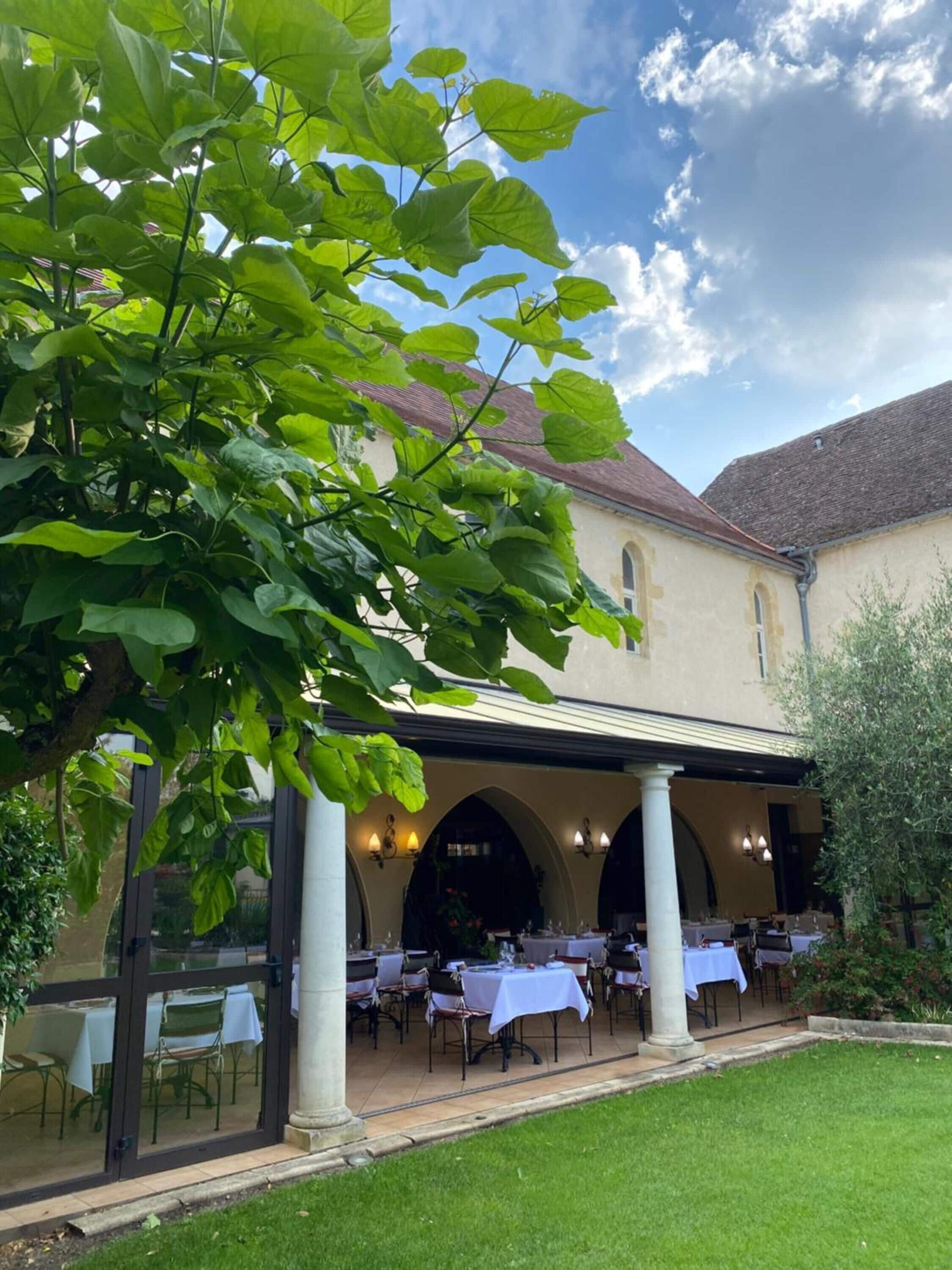 Un espacio de comedor al aire libre con mesas y sillas, un edificio con arcos y un árbol en primer plano.