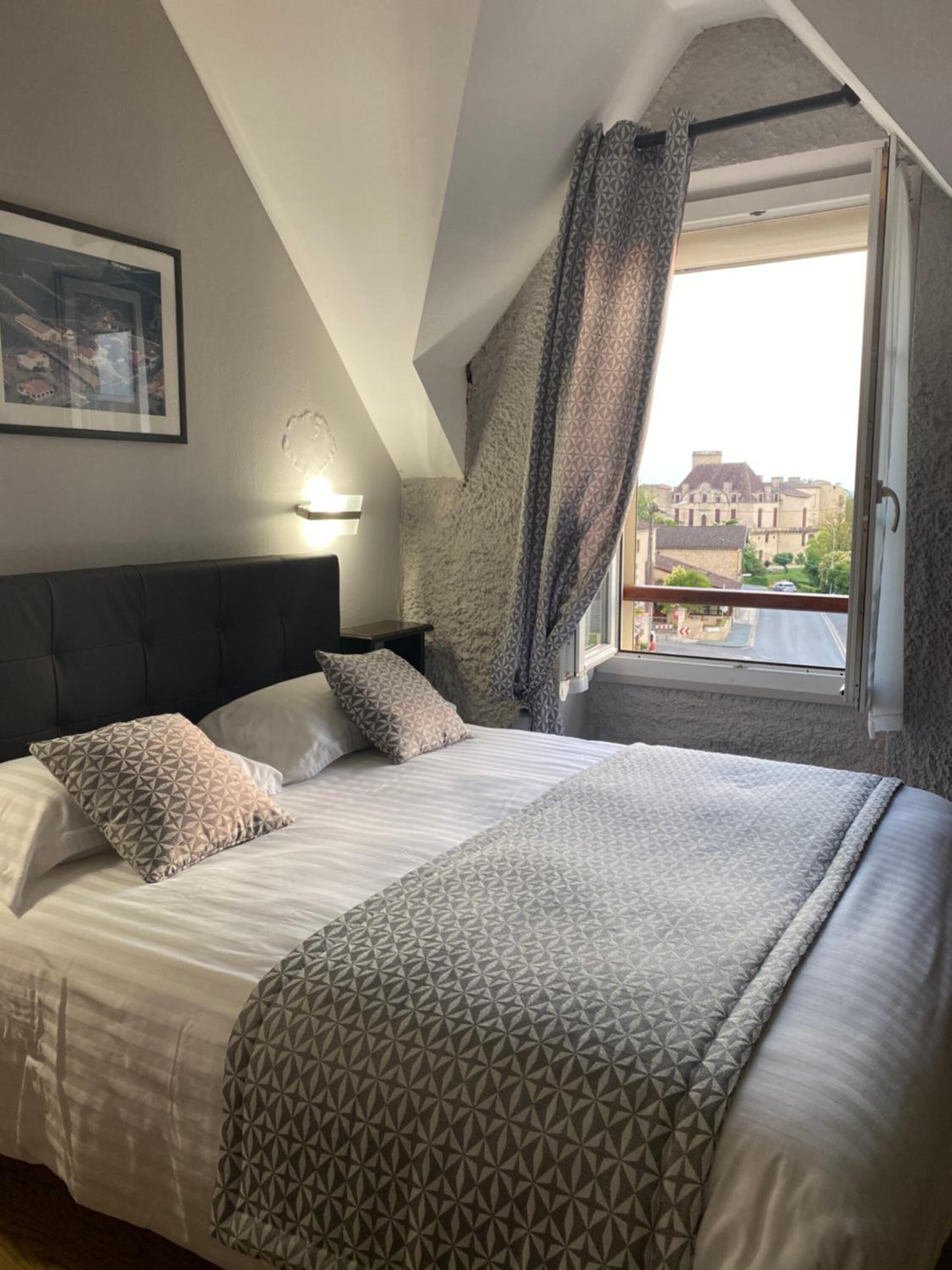 A bedroom with a large bed, a headboard, a nightstand, a wall-mounted light, and a window with curtains.