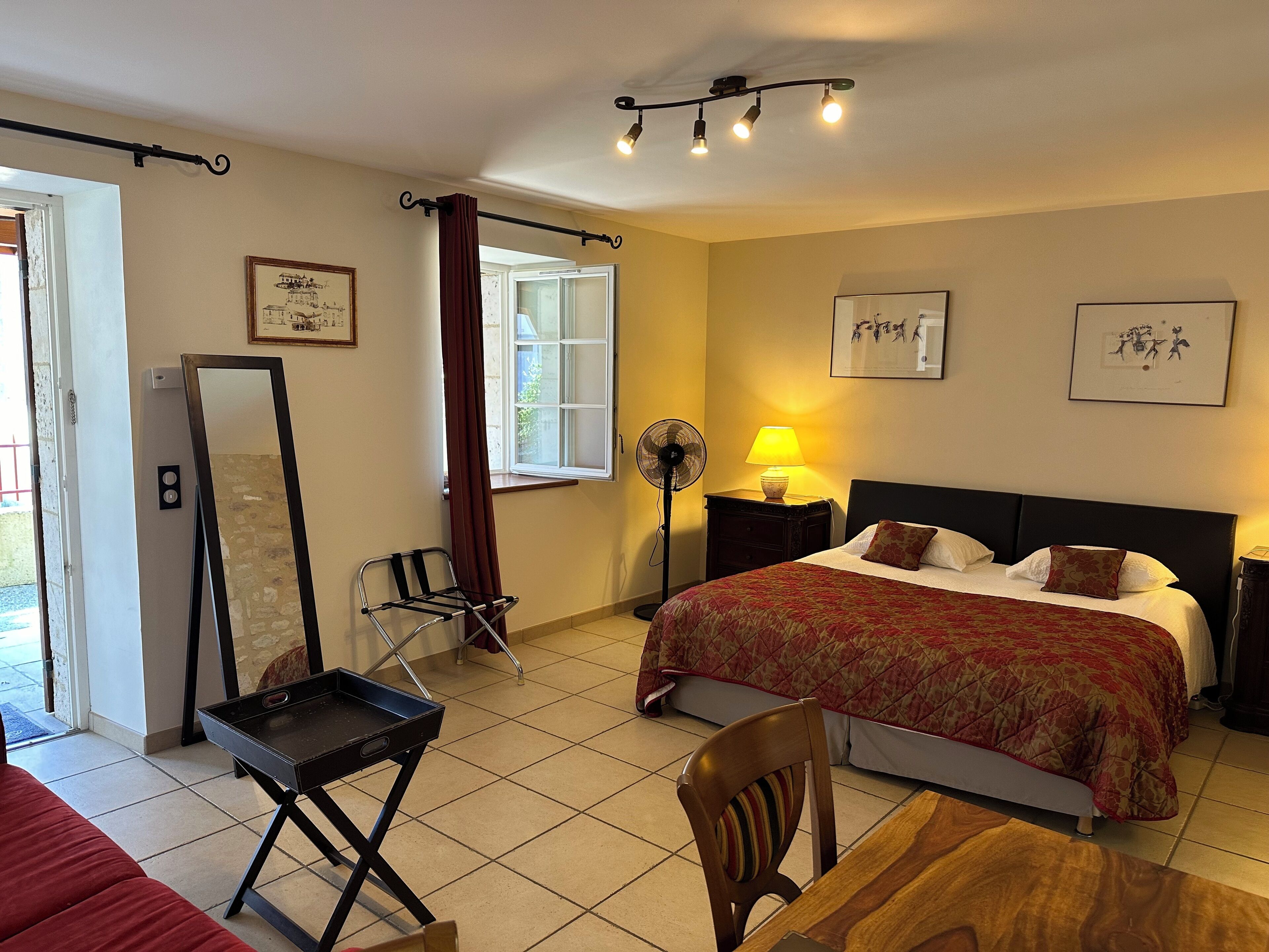 A hotel room with a bed, a desk, a chair, a standing fan, and a window with curtains.