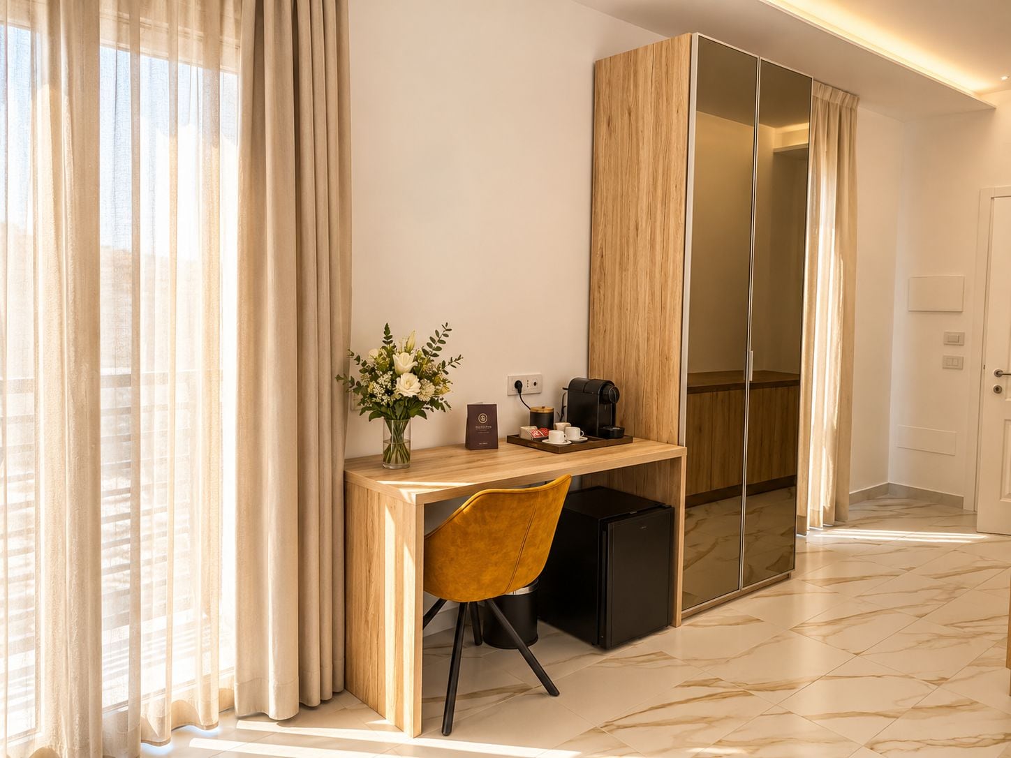 A modern hotel room with a wooden desk, a coffee machine, a vase with flowers, and a wardrobe with a mirror.