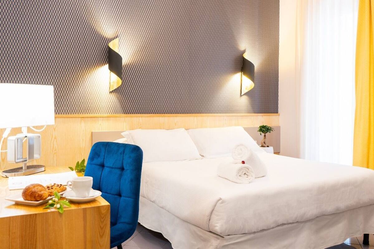 Une chambre d’hôtel avec un lit, une table de chevet avec une lampe, une chaise et une petite table sur laquelle se trouvent un croissant et une tasse.
