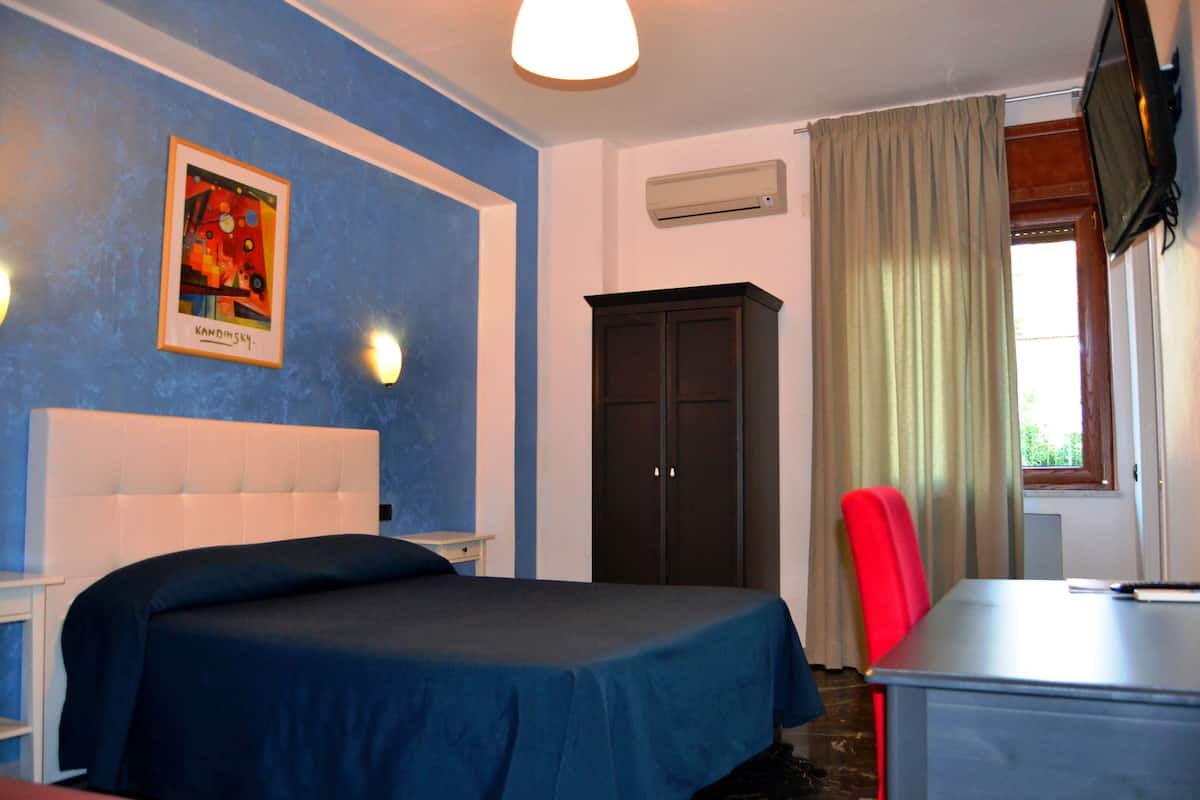 Une chambre à coucher avec un lit, un bureau, une chaise, une armoire, un tableau au mur et une fenêtre avec des rideaux.