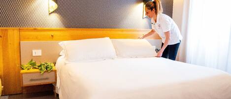 Una cama bien hecha en una habitación de hotel, con cabecera de madera, mesita de noche con planta y lámpara de pared.