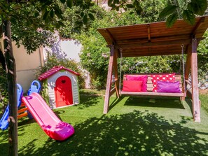 A wooden swing set with red cushions and a small slide in a backyard. - Diana Pompei (Pompei)