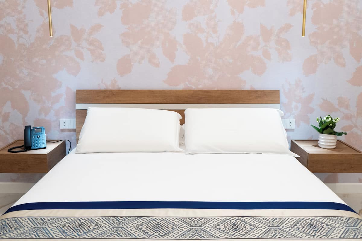 Un lit bien fait, avec une tête de lit en bois, deux tables de chevet sur lesquelles se trouvent un téléphone et un vase de fleurs, et une couvre-lit à motifs.