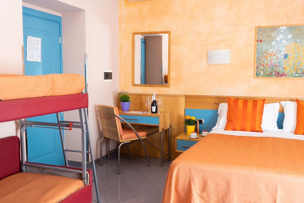 Une chambre d’hôtel avec des lits superposés, un bureau avec une chaise, une petite table avec une plante et des verres à vin, un miroir et un tableau floral accroché au mur.