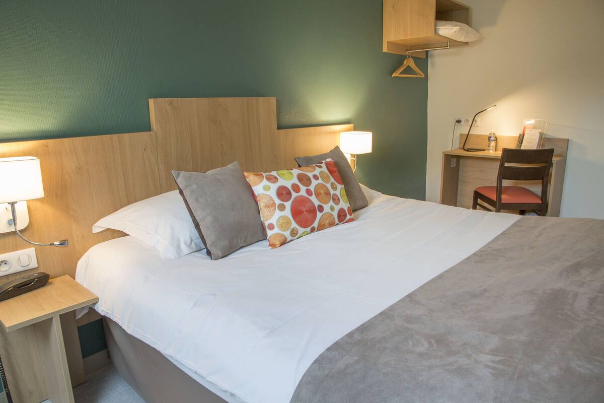A neatly made bed with a wooden headboard, a bedside table with a lamp, and a desk with a chair.